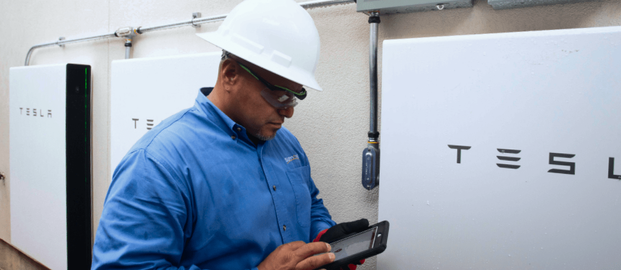 a man wearing a hard hat and a blue shirt standing in front of Tesla batteries. 